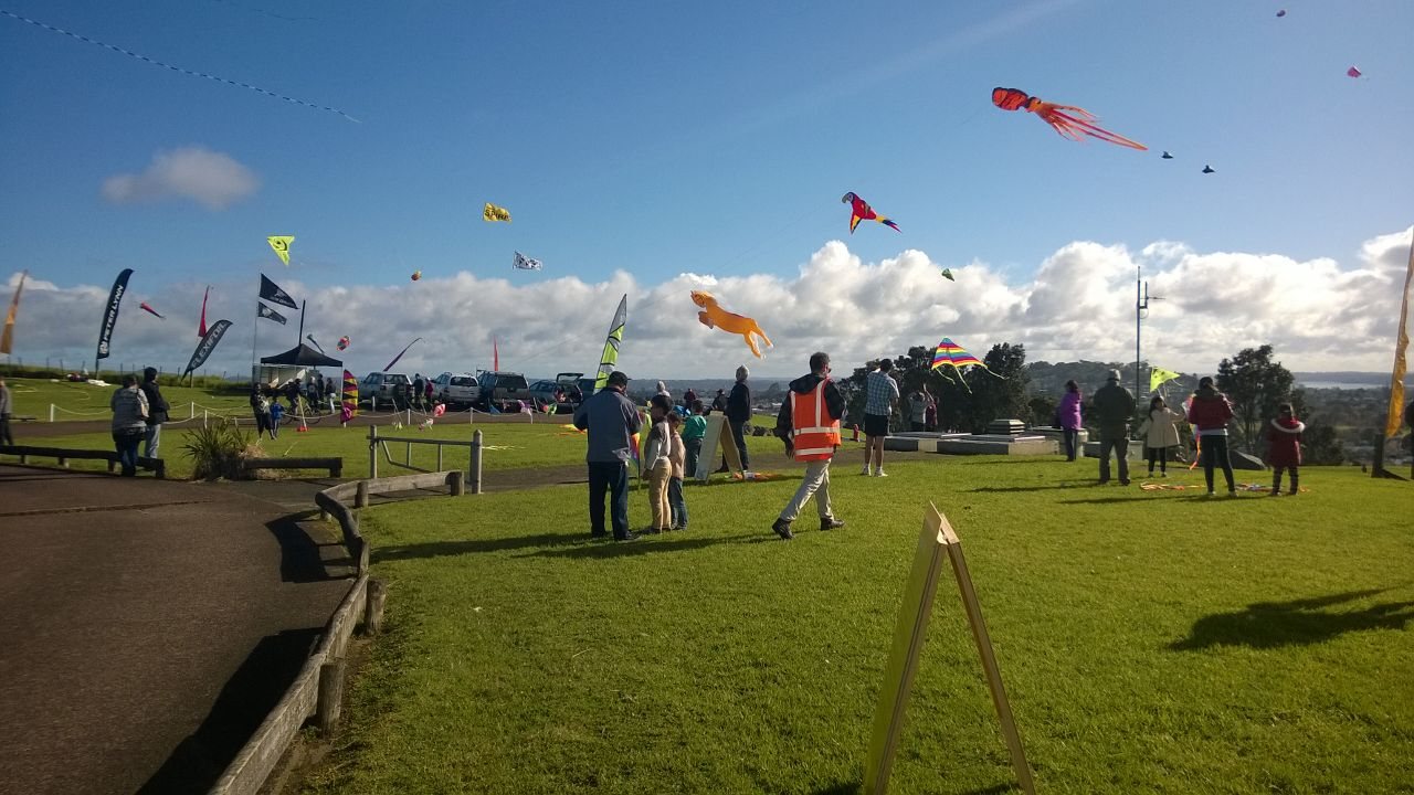 Families flying kites on the grassy summit of Mount Roskill under a blue sky with big white clouds